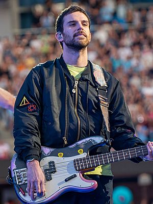 A short-haired man wearing a neon green shirt and a dark-coloured jacket plays the bass