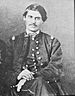 A white man with dark hair and a thin mustache sitting on a chair, his hands resting on his leg. He is wearing a military jacket and overcoat.