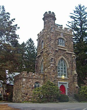 A three-story tower of stones in various shades of brown with a small flat-roofed projection on the left and a higher round turret on the front left corner, with tall evergreen trees behind it. It has arched windows, pointed on the first floor, with a red wooden door at the front.