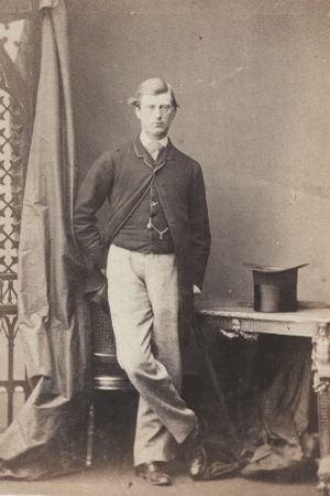 Standing photographic studio portrait of Ronald Leslie-Melville, wearing a jacket and waistcoat, with an inverted top hat on a table to his right