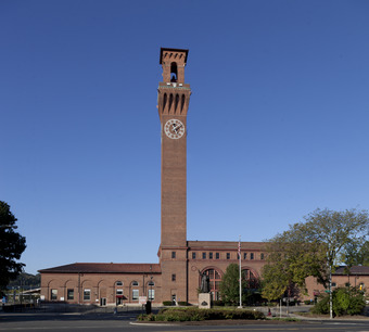 A brick building with a tall clock tower. Its two-story middle section has three tall round-arched windows and a peaked roof, with two one-story wings. In front is a square with a flagpole, statue and some small shrubbery and trees
