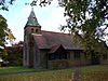 A small church with a tower and broach spire to the left, trees are to the left and overlapping to the front and autumn leaves lie on the grassed area in the foreground