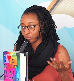 Woodfolk is holding a microphone and appears to be midsentence, gazing off-camera. She has brown skin, locs, and is wearing glasses and a black neck scarf over a red dress.