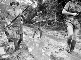 Three men in shorts, wearing steel helmets. Two carry rifles while the third has a submachinegun.