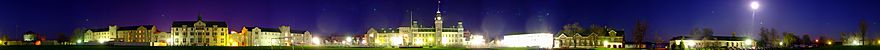 A 260-degree photo of the Royal Military College of Canada in Kingston, Ontario, on 4 May 2007. Seen is a green landscape during the night, featuring buildings made of white stone and red brick. The night sky is dark blue and purple, with the moon shining bright on the right side of the image. Photo credit: Martin St-Amant (User:S23678)