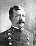 Head of a white man with neatly combed hair and a bushy mustache wearing a double-breasted military jacket with two columns of large buttons down the chest.