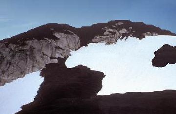 Rugged mountain covered with dark rock on its summit and flanks. Dark rock and patches of glacial ice loom in the foreground.