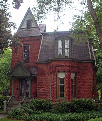 A brick house with a high pointed roof on its left and a lower, trapezoid-shaped one on its right. The first story has a pointed-roof porch on the left and a section sticking out from the brick on its right.
