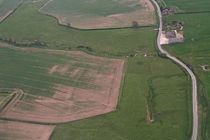 Aerial photograph of fields, with a less-than-straight country lane passing up  close to the right hand side.  In the top of the frame a modern farmhouse and buildings stand on right of the road. Most of the land is pasture, with two arable fields visible.  The crop there is sparse, with large bare patches.  The brook wiggles across the upper half of the picture, serpentine in a landscape of straight boundaries.  It is narrow and from this height and angle the water surface cannot be discerned.  The spring is centre right, its own water course straight and running upwards,toward the brook.  The spring is in the greenest of the meadows, with the low humps and bumps of the lost village around.