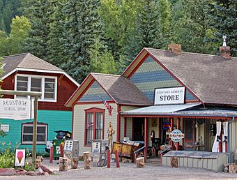 A colorful one-and-a-half-story wooden building with a pointed wooden shingled roof and large signs on the front and in front reading "Redstone General Store". There are some old gas pumps out front. To the left is another house in a mixture of colors.