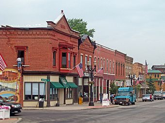 A row of buildings, mostly red and brown, with cars parked on the street next to them. The two on the left have pointy tops on their fronts, with the leftmost's having "1886" written in the middle. All the buildings have decorative window trim. There are American flags flying from the lampposts.