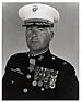 Head and shoulders of a middle-aged white man wearing a white peaked cap with oak leaf decorations on the visor and a dark military jacket with two rows of medals hanging from ribbons pinned to his chest and another medal hanging from his neck.