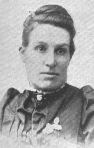 A white woman with dark hair dressed away from her face; she is wearing a dark dress with a high collar and puffed sleeves, with pins or ribbons on the chest