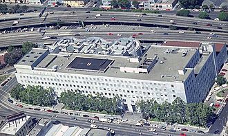 Aerial view of the San Francisco Hall of Justice building. It is a multistory building that occupies the entire length of a city block along Bryant Street, between 7th and Harriet; the Dwight D. Eisenhower Freeway (Interstate 80) has its western terminus near and curves around the back of the building. There is a prominent black asphalt helipad on the roof.