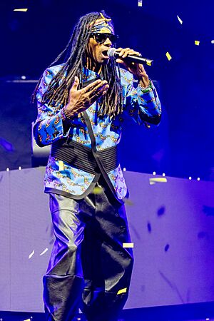 Fab Morvan wearing black leather pants, a colourful, patterned jacket, and black sunglasses, singing into a microphone onstage