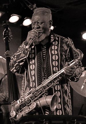 Sanders holding a microphone with a saxophone slung over his body