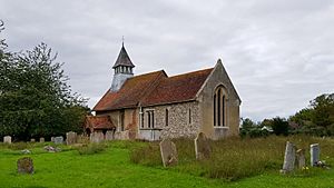 A small stone church with red tiled roofs seen through a churchyard from the south-east.  On its far gable is a bellcote with a pyramidal roof