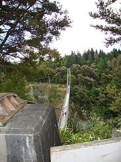 View of suspension bridge, with the foundation for the suspension cables in the foreground and a steel lattice tower in the background, amidst the forested gorge.