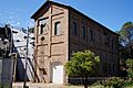 Photograph of the Folsom Powerhouse, a tall, narrow masonry building with power lines overhead and the dam in the background.