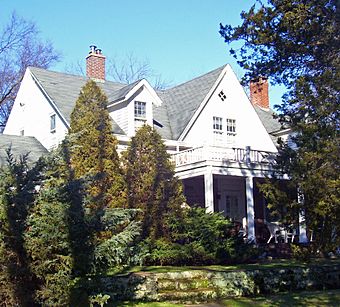 A white house with pointed gray roofs and a front porch behind evergreen trees and shrubs. There is a stone retaining wall in front, at the bottom of the image.