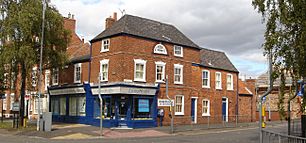 The corner of a terraced suburban street. The lower storey is a corner shop, now advertising as a chiropractic clinic. The building is two storeys high, with some parts three storeys high. It was formerly Alfred Roberts' shop.