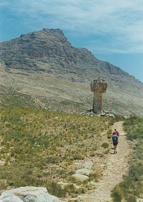 Hikers walking a path towards a rock pillar; in the background a steeply-sloping mountain.
