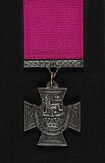 A bronze cross pattée bearing the crown of Saint Edward surmounted by a lion with the inscription "for valour". A crimson ribbon is attached