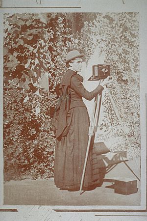 Greyscale image of Emma Jane Gay (known as E. Jane Gay), with her camera and tripod. The woman is wearing a long dress and a bowler hat, and the background depicts walls covered in ivy.