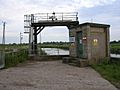 A metal gantry between the road in the foreground and a river. To the right is a breeze block building with warning signs on it.