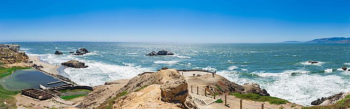 Panorama shows a wide, sweeping view of the view from an outcropping at Land's End.  The Pacific Ocean shines a brilliant aquamarine and spectators can be see enjoying the ocean breeze on the clear summer day.
