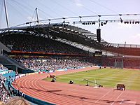 A fully occupied grandstand on a sunny day. In front of it is an athletics track.