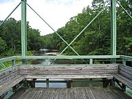 A detail of the wooden seat and observation platform at the center of the green Whipple truss bridge facing out over a river
