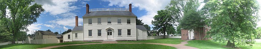 A two-story white house with one-storey wings on each side, many windows, a central door, three chimneys, and a balustrade. A white latrine and barn are on the left, and a small red brick building with a white door is on the right. A white wooden fence encloses the whole yard, which has several large trees. The sky is blue with white clouds.