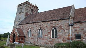 The nave of a stone church seen from the southwest, containing three pointed windows and a small porch, with a battlemented tower beyond