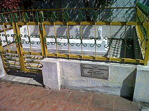 Photograph of a white tomb surrounded by a low white wall topped with a gold coloured fence and with a plaque on the wall to the right of the entrance