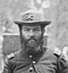 Head of a white man with a full bushy beard, wearing a cavalry hat and a military jacket with a single button buttoned near the neck.