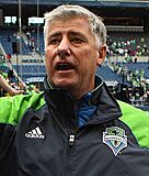 A headshot of Sigi Schmid wearing a Seattle Sounders FC tracksuit, pictured at a stadium