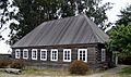 Photograph of the Fort Ross Commander's House on a sunny day. Visitors stand at a table in front of the rectangular log building with a high, peaked roof and white painted windowframes.