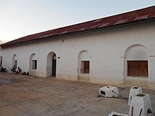 The white facade of a single-storey building, extending away from the viewer to the left. There is a single arched doorway, and a row of windows set into arched recesses. The roof is rusty corrugated metal. Four low white posts are linked by chains in the right foreground, one of them has been knocked over. Two ladies are visible in front of the left-most window. One sits on a dark plastic chair looking into a pram, the other stands behind the pram, also looking at it.