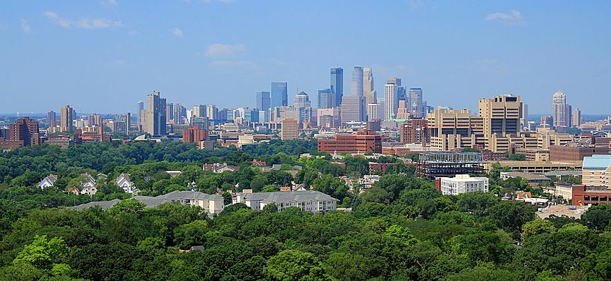 The Minneapolis skyline rises to its highest point at the center of the image, with the three tallest buildings standing out against a clear blue sky. Before the skyline are trees, university buildings, and residential complexes.