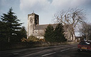 A church with a red tiled roof and a square tower. Trees in the foreground partially obscure the building. The sky is overcast and grey.