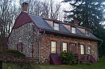 A small house made of reddish stone. It has a curved roof with three windows. Near the top of the roof the house's siding is maroon-colored wood.