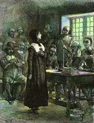 A woman standing before a table behind which are seated several men, with several other men occupying seats against the walls of the room