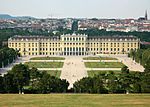 Rococo three and four story palace stretches across most of the midground. In the foreground are manacured lawns and walkways, while the background is the old city of Vienna with a cathedral on the horizon.