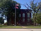 A dark red brick rectangular two and a half-story courthouse hidden behind large trees with a tall flagpole in the front yard