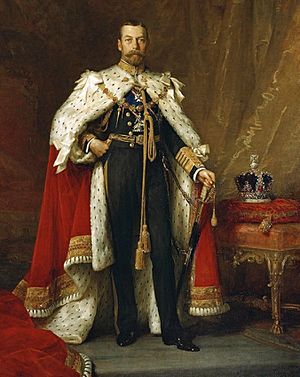 Full-length portrait in oils of King George V of the United Kingdom. He wears naval uniform under an ermine cape, and beside him a jewelled crown stands on a table.