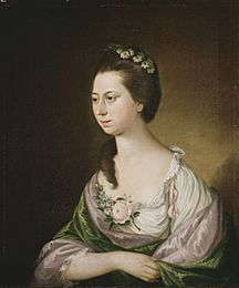 Portrait of a young woman at half length. She wears a cream top with a green shawl, and has a floral posy over her cleavage. She has flowers in her hair, which is long but swept back to expose her neck.