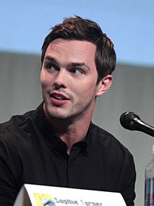 A young, Caucasian man with short, dark hair and facial stubble wearing a black shirt speaks into a microphone against a grey and blue background.