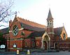 Three-quarter view of an ornate brick building with pale stone dressings and recessed lancet and round windows. A rose window dominates the left-hand side.  Perpendicular to its right, there is an arched entrance porch; beyond that is an octagonal tower with louvre-style openings below a conical spire.