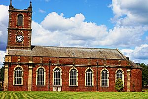 A brick church with stone dressings seen from the south.  The west tower has a clock and pinnacles, and along the south face of the body of the church are Georgian-style windows.